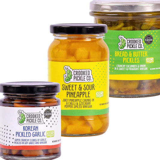 CROOKED PICKLE CO The OG Pickle Trio with a variety of pickles in glass jars arranged on a wooden surface.