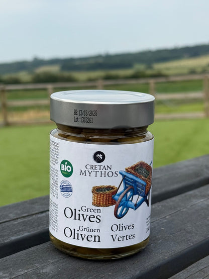 Organic Greek green olives in brine from Cretan Mythos, displayed in a clear jar with olives and herbs visible inside.