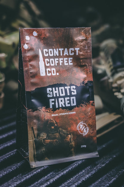 A cup of Shots Fired Coffee from Contact Coffee Company Ltd. with a rich, dark brew and branded packaging in the background.
