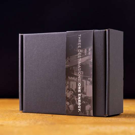 A dark charcoal gray rectangular product box with a vertical black-and-white printed label reading 'THREE DESTINATIONS ONE ENERGY,' photographed on a wooden surface against a dark background.
