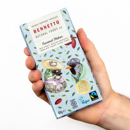 Hands holding Bennetto Natural Foods UK chocolate coconut flakes, close-up of shredded coconut covered in rich chocolate
