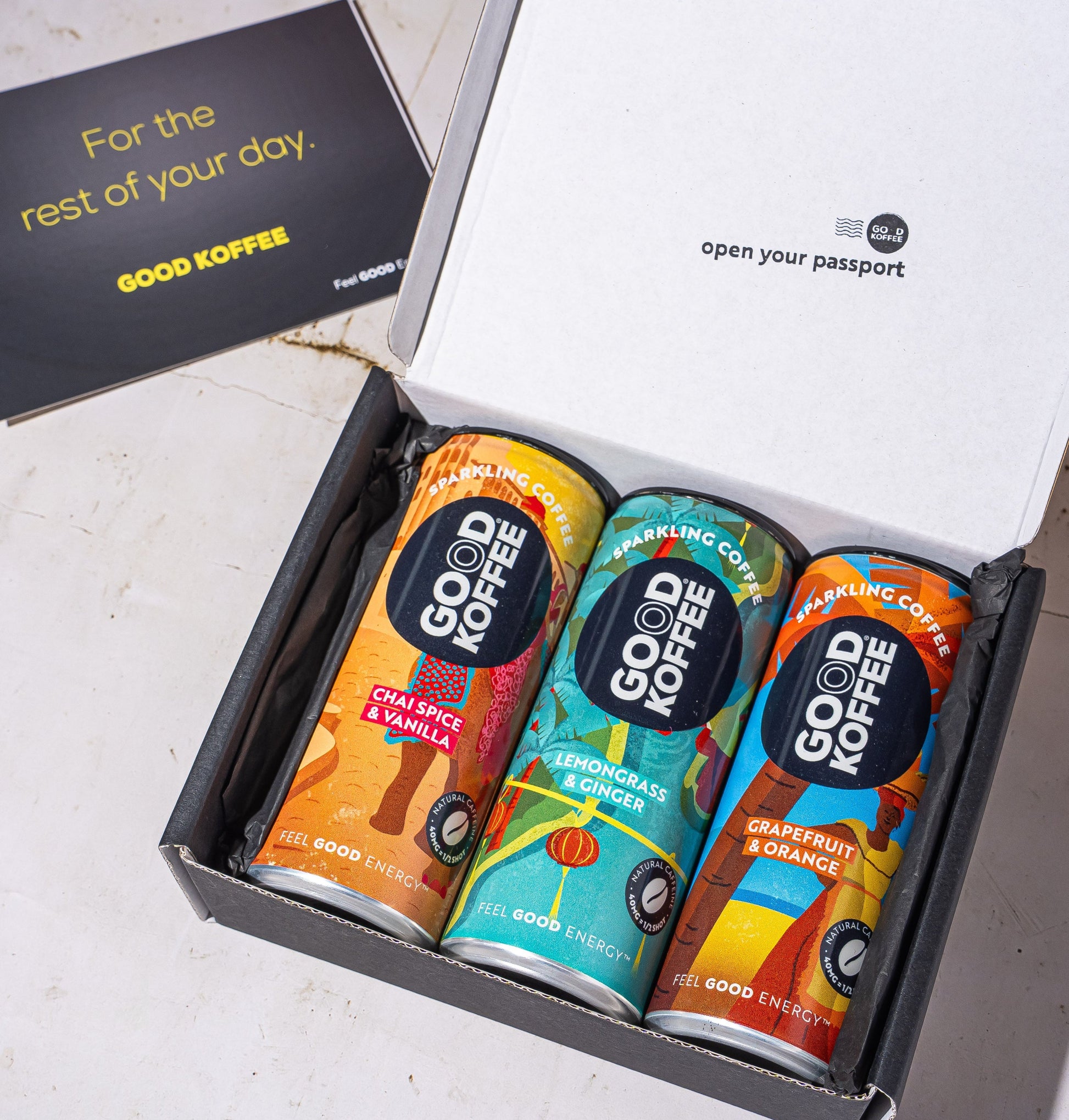 An open black gift box containing three colorful Good Coffee cans, with a black card beside it on a light stone surface. The inside lid reads 'open your passport' and the card says 'For the rest of your day.