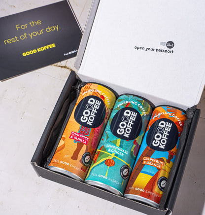 An open black gift box containing three colorful Good Coffee cans, with a black card beside it on a light stone surface. The inside lid reads 'open your passport' and the card says 'For the rest of your day.