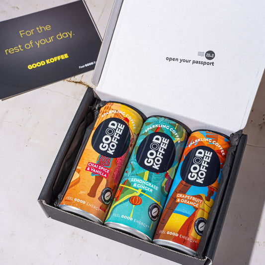 An open black gift box containing three colorful Good Coffee cans, with a black card beside it on a light stone surface. The inside lid reads 'open your passport' and the card says 'For the rest of your day.