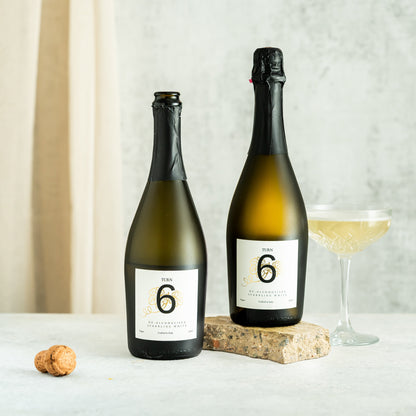 Six bottles of Turn 6 premium alcohol-free sparkling white wine in a case, showing clear glass bottles with white wine and foil caps.