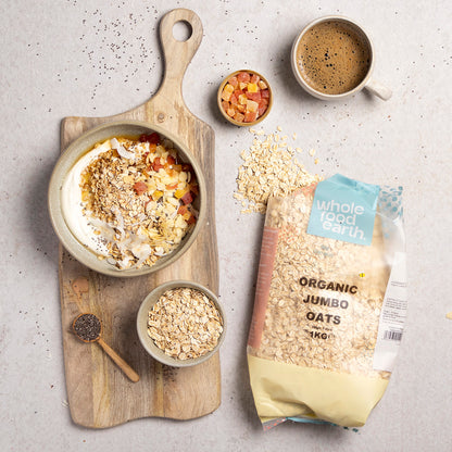Bowl of organic jumbo oats from Whole Food Earth with a spoon, placed on a wooden table with fresh fruits and a jar in the background.