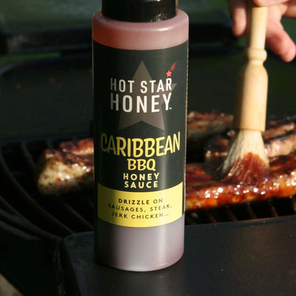 Close-up of Hot Star Honey Caribbean BBQ sauce bottle with barbecue grill and grilled meats in the background.