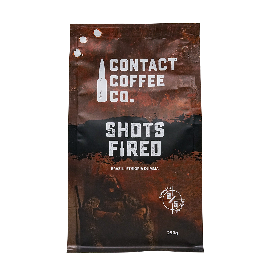 A cup of Shots Fired Coffee from Contact Coffee Company Ltd., showing rich coffee with a frothy top and branded packaging.