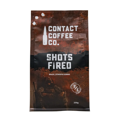 A cup of Shots Fired Coffee from Contact Coffee Company Ltd., showing rich coffee with a frothy top and branded packaging.