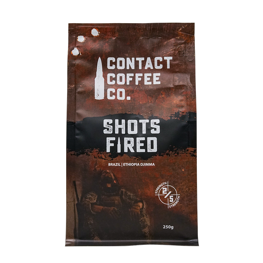 A cup of Shots Fired Coffee from Contact Coffee Company Ltd., showing rich coffee with a frothy top and branded packaging.