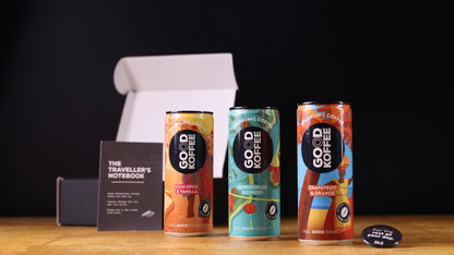 Three cans of GOOD KOFFEEcsparkling coffee displayed on a wooden surface with a black background, alongside a black box, a traveller's notebook, and a small round button badge.