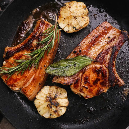 Two cooked Primrose Herd pork chops sizzling in a pan, ready to serve on a dinner plate.