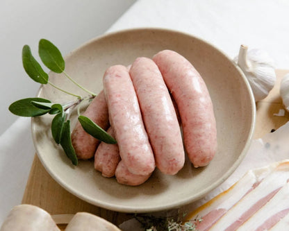 Primrose Herd pork sausages, Cumberland style, shown raw on a wooden cutting board with herbs and seasoning.