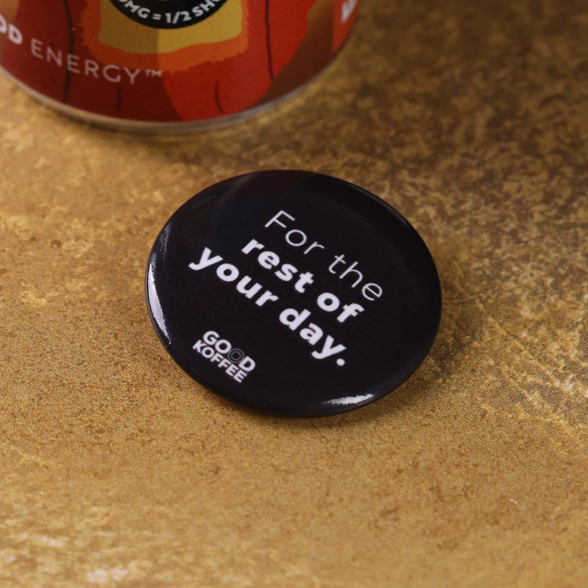 A round black coffee-themed pinback badge with the white text 'For the rest of your day.' and 'GOOD KOFFEE' logo, resting on a textured brown surface with a coffee can partially visible in the background.