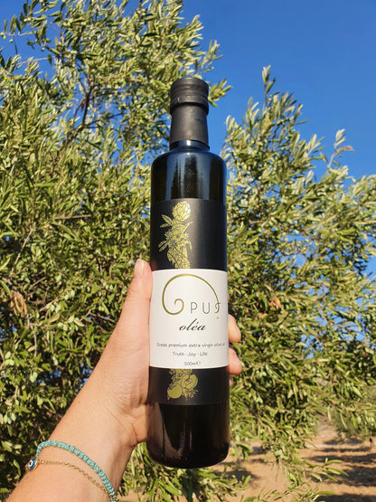 Opus oléa extra virgin olive oil 500ml bottle in a grove setting with olive branches and trees in the background.