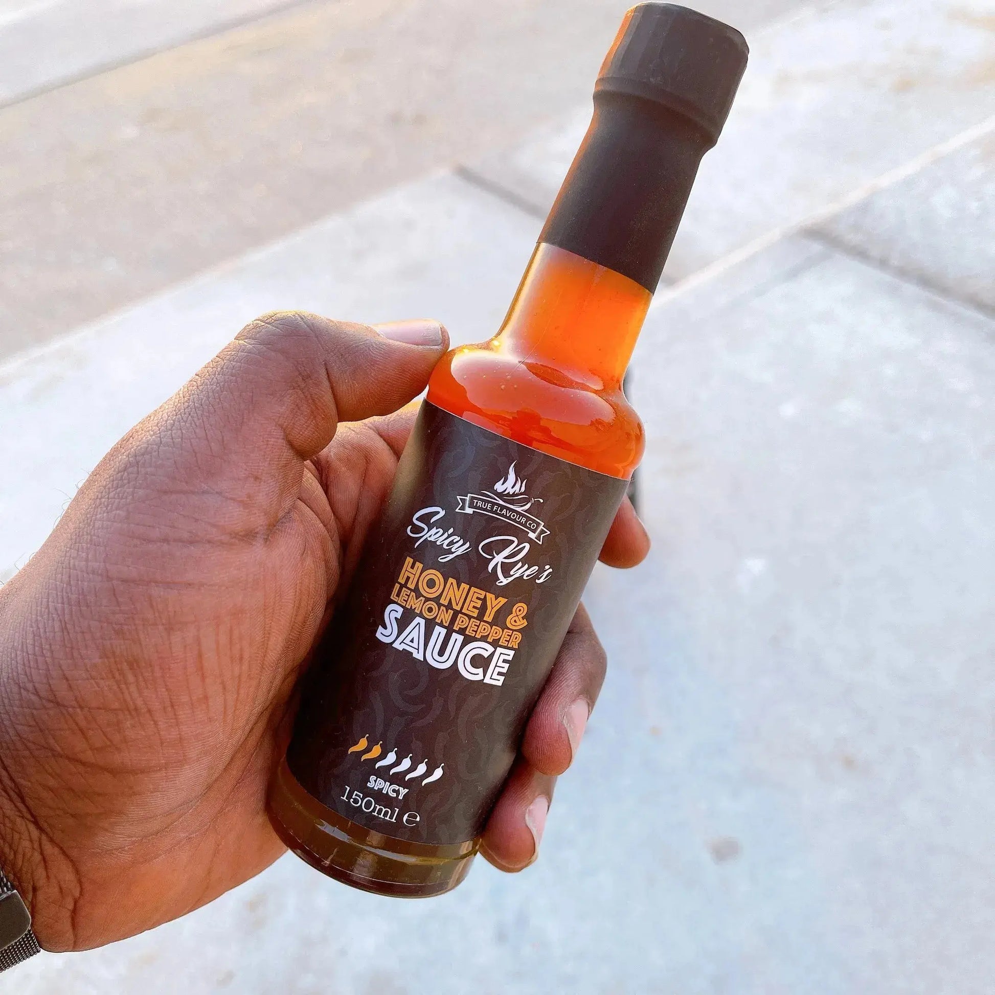 Spicy Ryes Honey & Lemon Pepper Hot Sauce bottle with yellow lemon slices and black peppercorns on a wooden surface.
