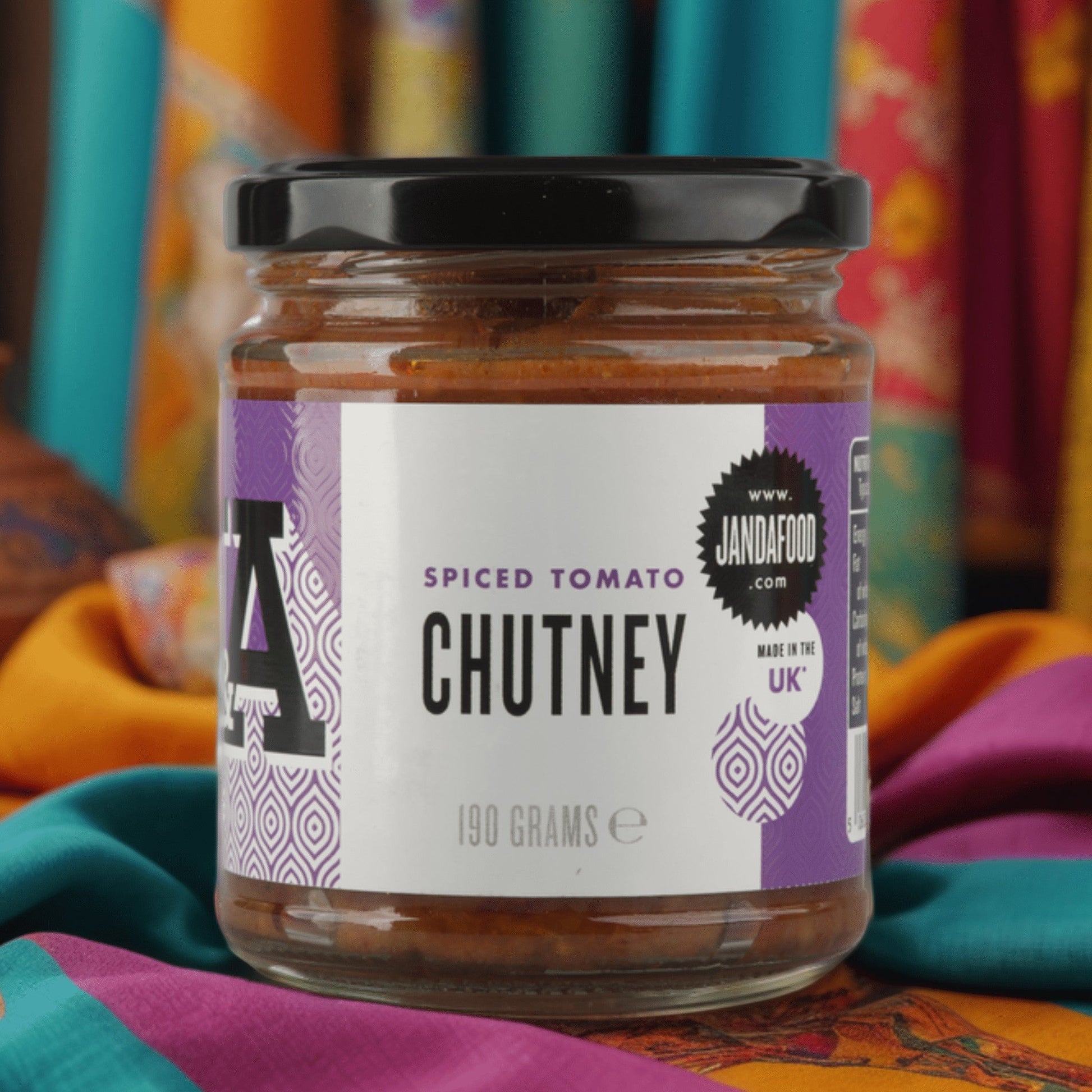 Janda Food Spiced Tomato Chutney in a glass jar with a purple label, set against a vibrant patterned fabric backdrop.