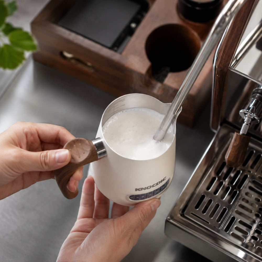 KNOCDIC® milk frothing pitcher with built-in thermometer, shown in use for steaming milk for coffee brewing.
