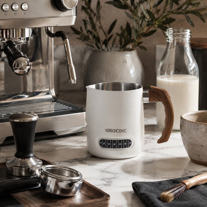 KNOCDIC® milk frothing pitcher with built-in thermometer for precise steaming, shown in a barista setup with frothing tools.