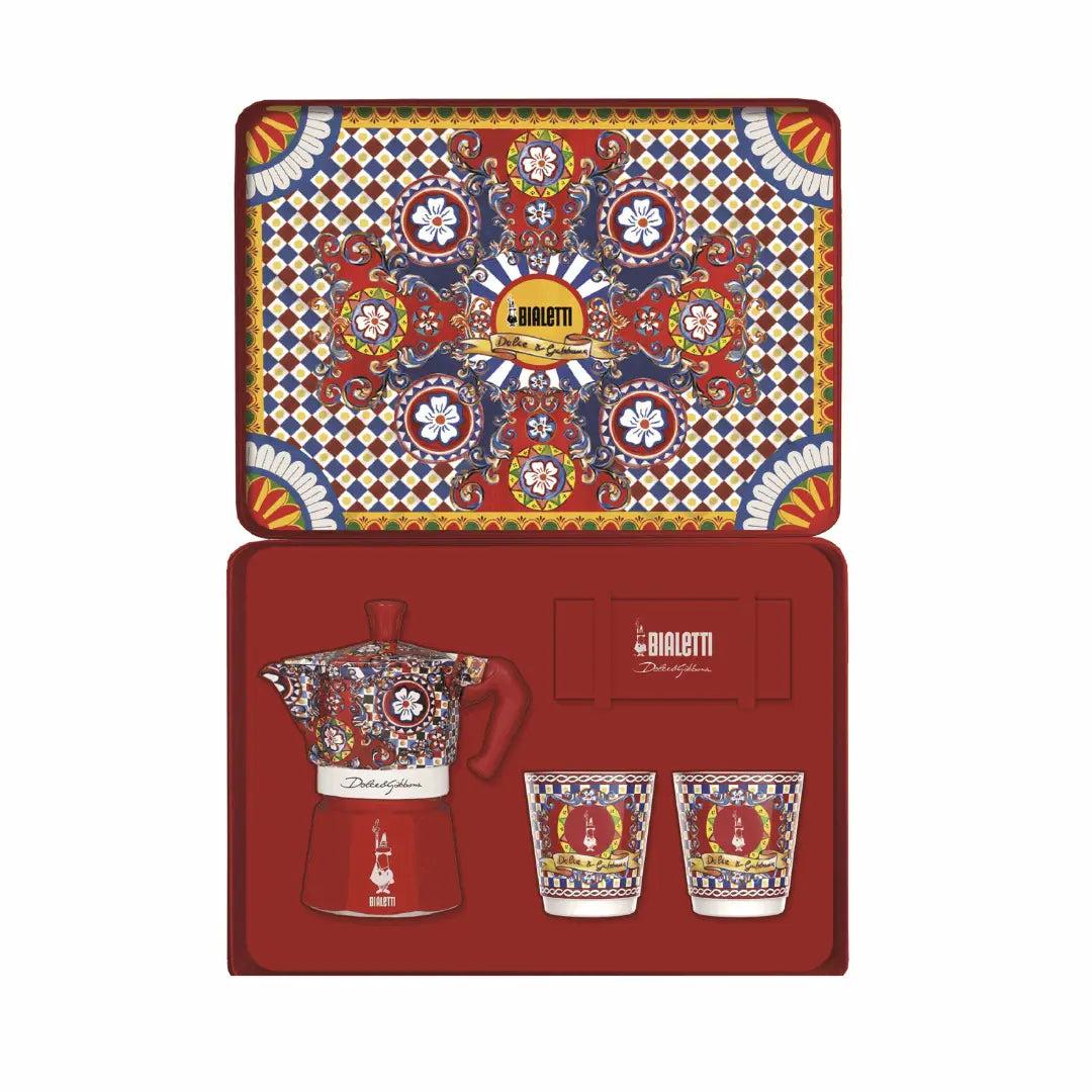 Bialetti X Dolce&Gabbana Sicilian Cart gift set includes 2-cup moka espresso maker, 2 cups, and gold stirrers on festive background.