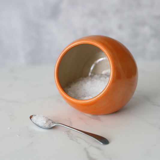 Orange ceramic salt pig with a matching spoon, designed by Katie L Thompson for easy salt storage and serving.